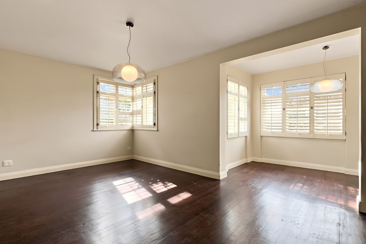 A bright, empty living area featuring dark hardwood flooring, neutral walls, and plantation shutters on the windows.