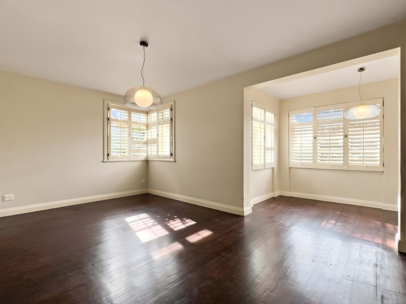 A bright, empty living area featuring dark hardwood flooring, neutral walls, and plantation shutters on the windows.