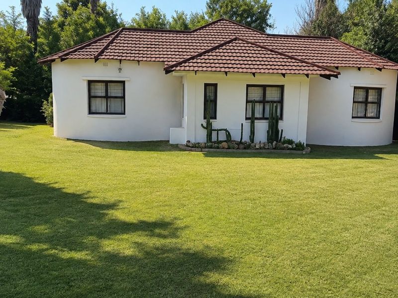 A well-maintained single-story house featuring a tiled roof, white exterior walls, and large windows, set within a spacious, manicured garden.
