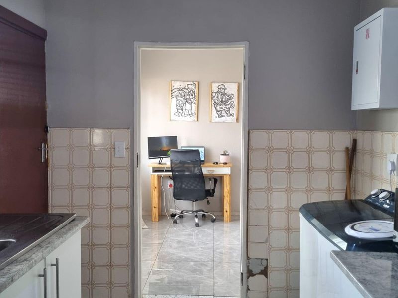 A functional kitchen area featuring built-in cabinetry, stone-look countertops, and tiled splashbacks, leading into an adjacent office space.