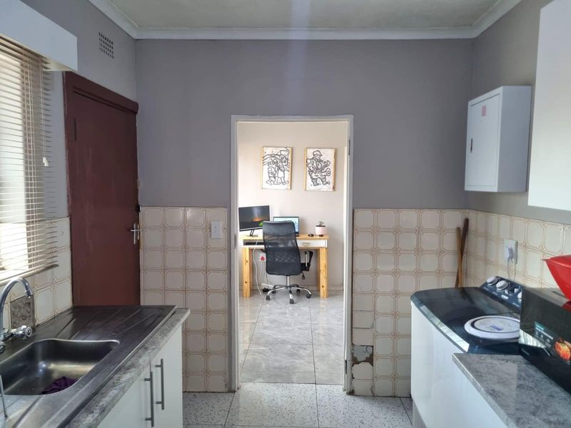 A functional kitchen featuring tiled splashbacks, built-in cabinetry, and a stainless steel sink, with a doorway leading to an adjacent office space.