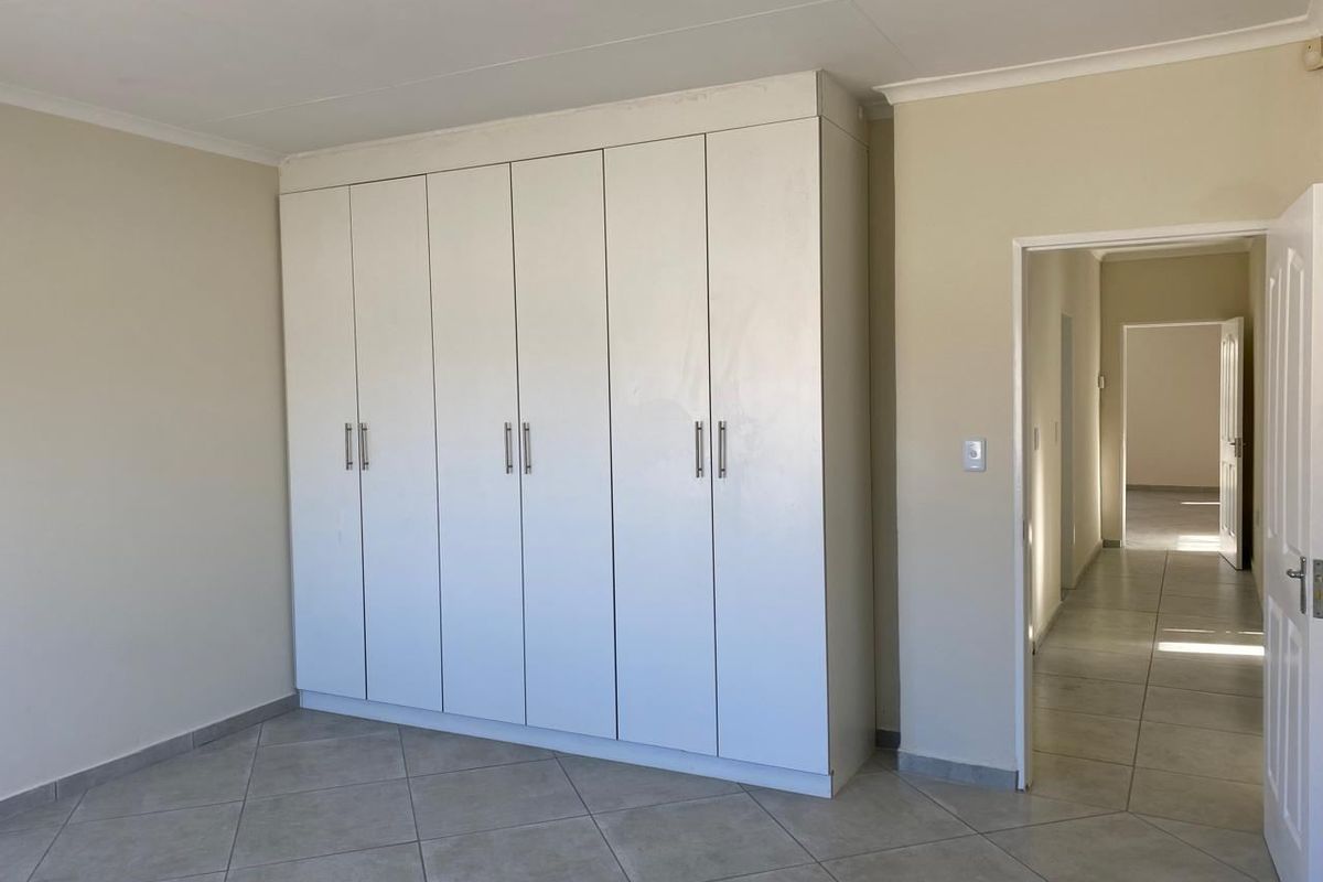 A clean, empty bedroom featuring neutral tiled flooring and ample built-in cupboard storage, leading into a bright hallway.