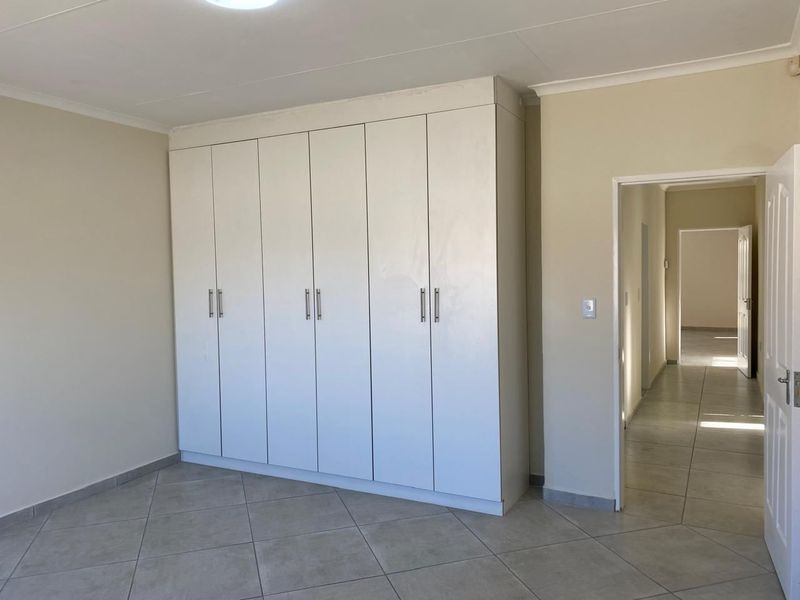 A clean, empty bedroom featuring neutral tiled flooring and ample built-in cupboard storage, leading into a bright hallway.