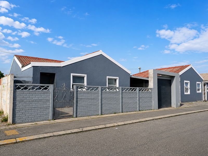 Front exterior view of a single-story house featuring a grey painted facade, tiled roof, secure boundary wall with gate, and a roller garage door.