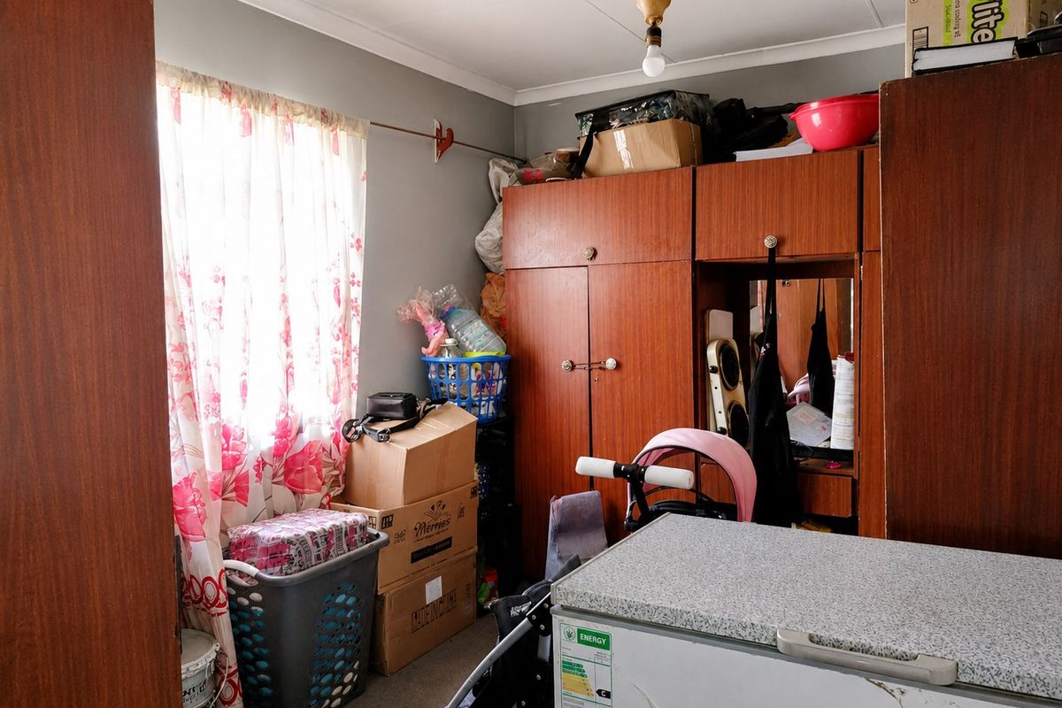 A bedroom featuring built-in wooden cupboards and a window with curtaining, currently used for storage.