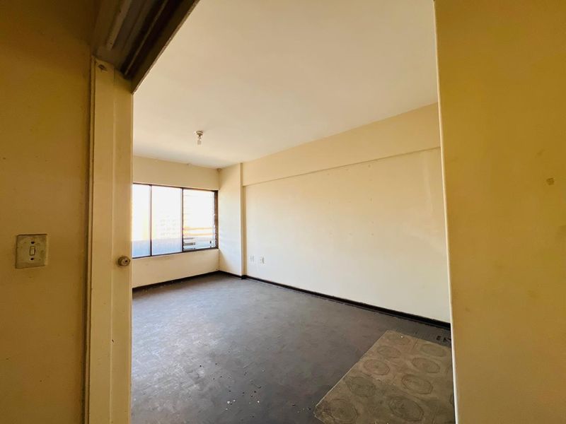 An empty, dated bedroom featuring plain walls, worn flooring, and an aluminium-framed window, offering a blank canvas for renovation.