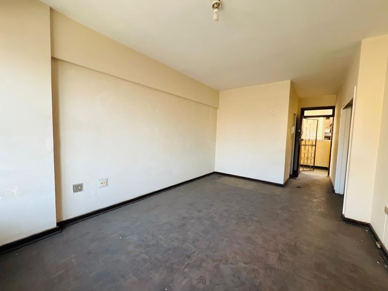 An empty, neutrally painted living area featuring worn flooring and direct access to an adjacent room through a security gate.