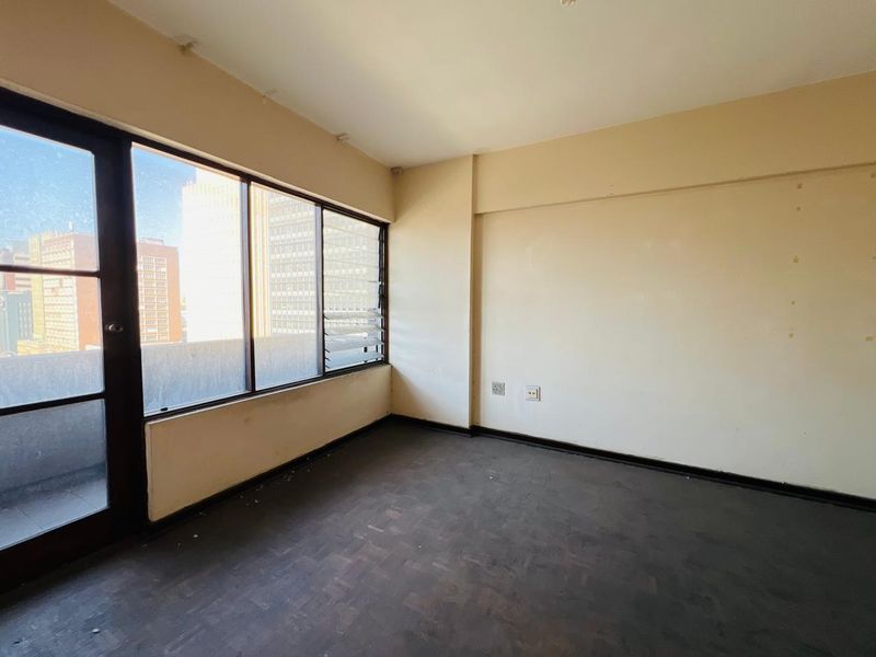 An empty bedroom featuring dark parquet flooring, large aluminium-framed windows with louvered sections, and access to an adjacent balcony area.