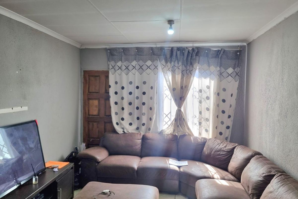 A living room featuring tiled flooring, a timber door, and security gate, with visible wear on the wall finishes and ceiling panels.