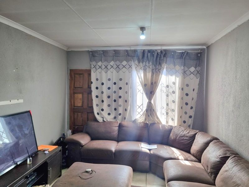 A living room featuring tiled flooring, a timber door, and security gate, with visible wear on the wall finishes and ceiling panels.