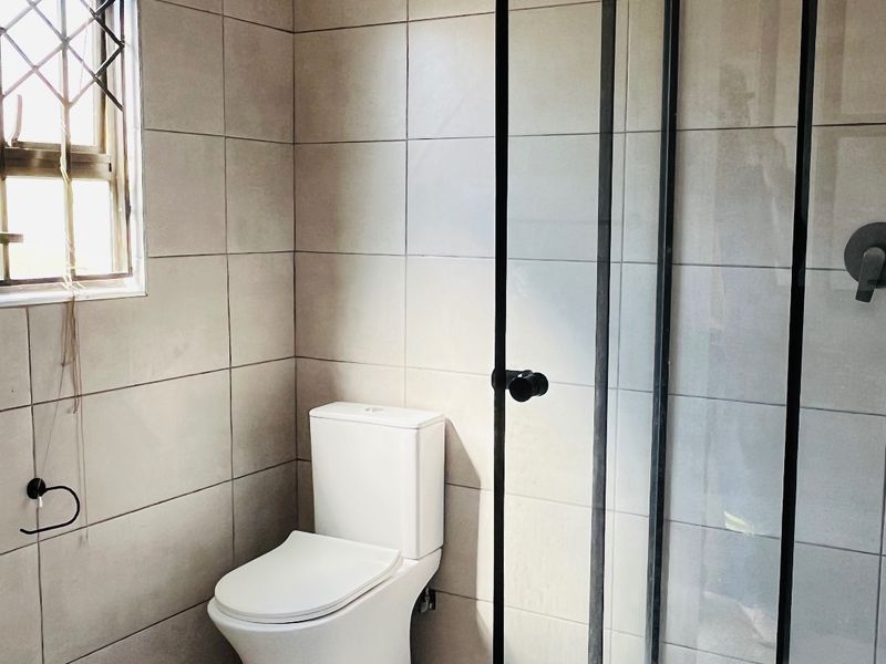 A clean, modern bathroom featuring a glass-enclosed shower, neutral wall and floor tiling, and a standard toilet.