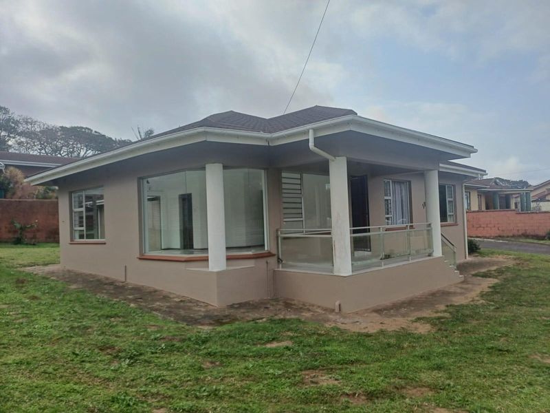 A well-maintained single-story house featuring a covered front patio with glass balustrades, concrete columns, and large corner windows set on a spacious lawn.