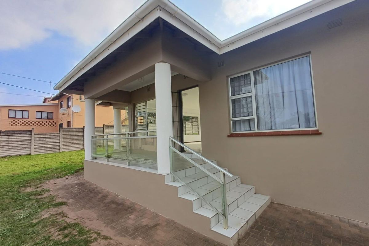 A well-maintained rear exterior view featuring a covered tiled patio with glass balustrades, concrete columns, and paved access steps.