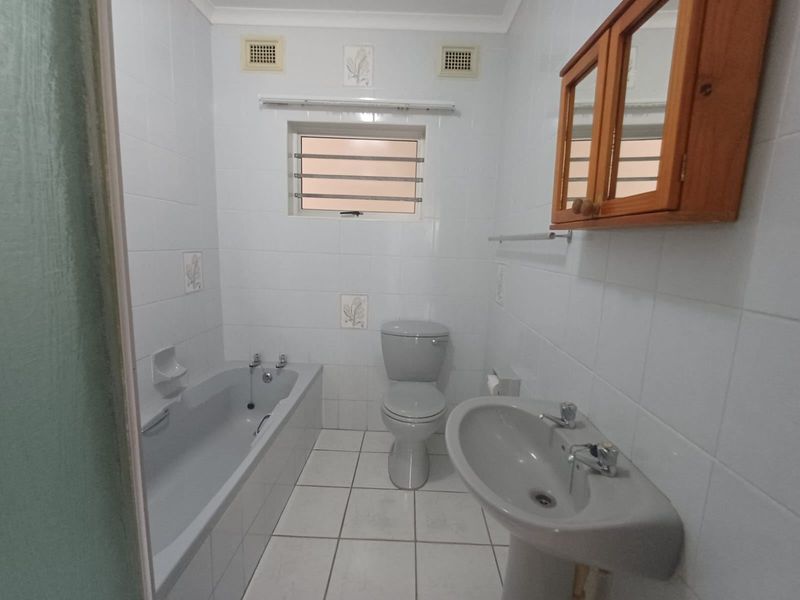 A functional bathroom featuring a bathtub, pedestal basin, and toilet with white wall tiling and louvered window ventilation.