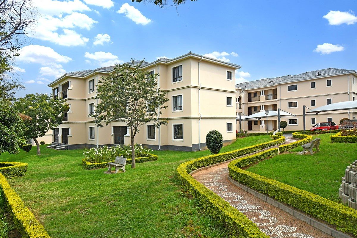 A well-maintained apartment complex featuring landscaped gardens, paved walkways, and shaded parking areas within a peaceful residential estate setting.