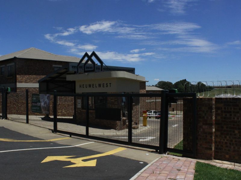 The entrance to Heuwelwest estate featuring a secure gated access point, brick perimeter walls with razor wire, and a dedicated security guard booth.