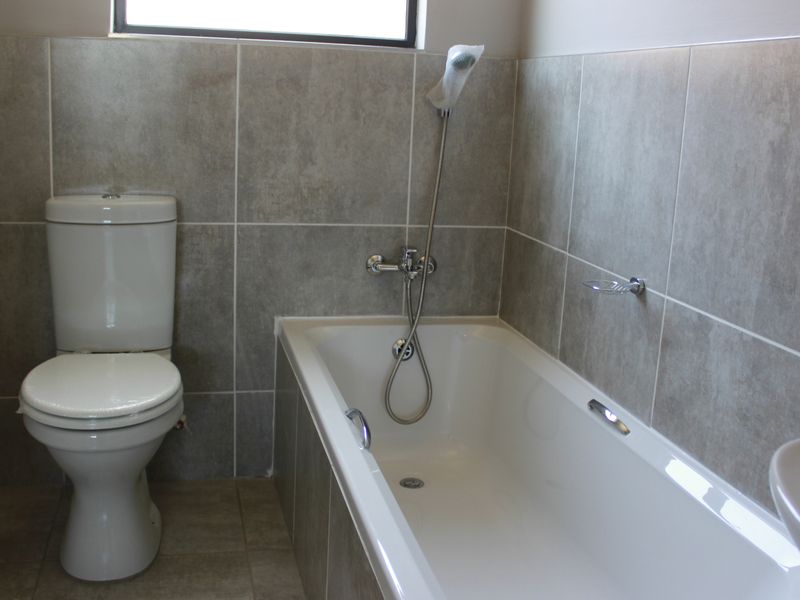 A clean, modern bathroom featuring neutral grey wall tiles, a white bathtub with a handheld shower mixer, and a standard toilet.