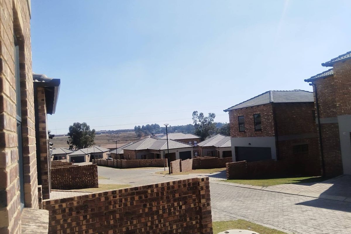 A view of a residential street within a housing estate, featuring face brick homes, paved driveways, and boundary walls under a clear sky.