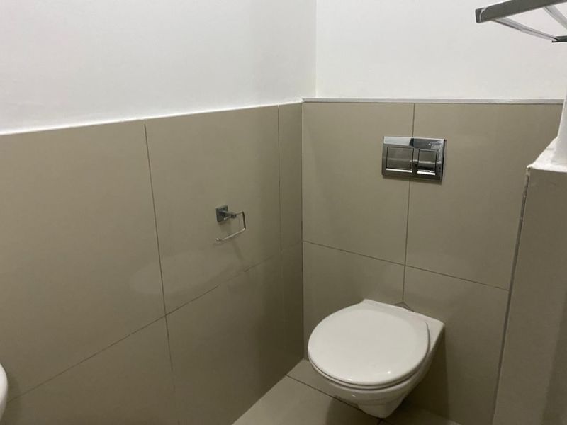 A clean and functional bathroom featuring a wall-mounted toilet with a chrome flush plate and neutral-toned wall tiling.