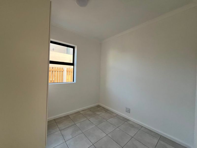 A clean, empty bedroom featuring neutral tiled flooring, a built-in cupboard, and a window with an aluminium frame.