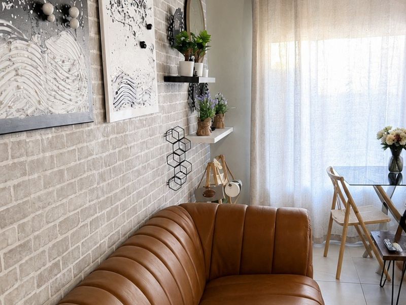 A cozy living area featuring textured faux brick wall finishes, neutral floor tiles, and natural light filtering through window curtains.
