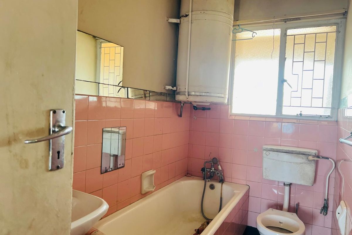 A dated bathroom featuring original pink wall tiles, a pedestal basin, a bathtub with shower attachment, and a wall-mounted geyser.