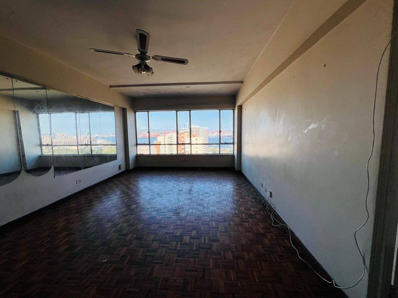 An empty living room featuring parquet flooring, a ceiling fan, and a large wall mirror, offering expansive city and harbor views through large windows.