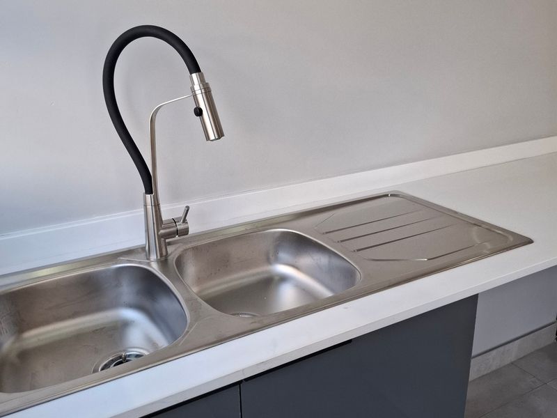 A clean, modern kitchen area featuring a stainless steel double sink with a contemporary gooseneck faucet, set against grey cabinetry and a white countertop.
