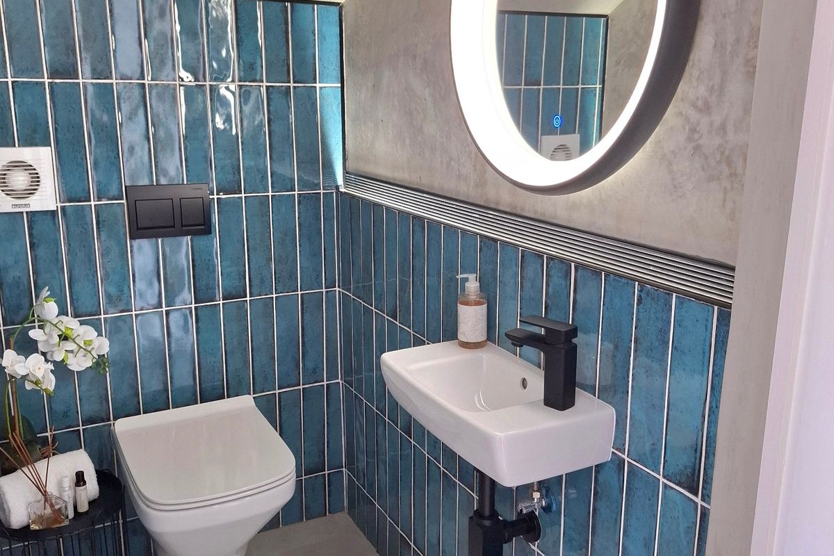 Modern bathroom featuring striking blue vertical wall tiles, a wall-mounted basin with black fixtures, a wall-hung toilet, and a contemporary backlit circular mirror.