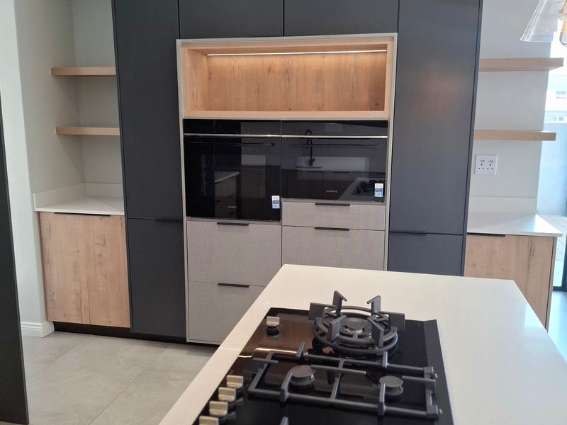 Modern kitchen featuring dark cabinetry, light wood accents, stone countertops, a central island with a gas hob, and built-in ovens.