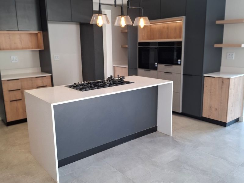 Modern kitchen featuring a central island with a gas hob, built-in ovens, sleek dark cabinetry with wood accents, and large format floor tiles.
