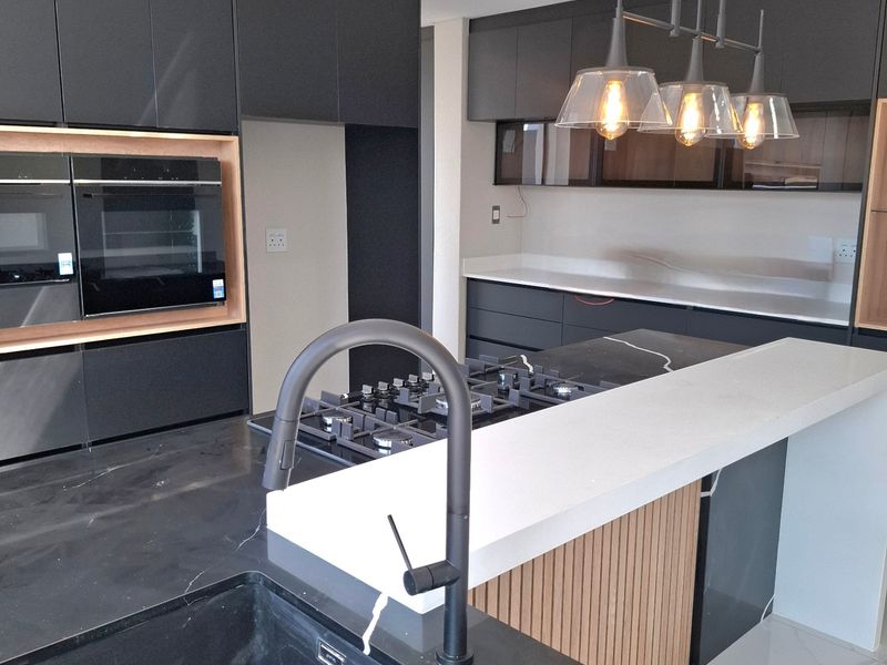 Modern kitchen featuring dark cabinetry, stone countertops, a central island with a gas hob, integrated ovens, and high-end finishes throughout.