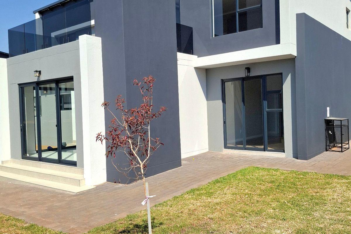 Rear view of a modern two-story house featuring a paved patio, glass sliding doors, and a glass-railed balcony.