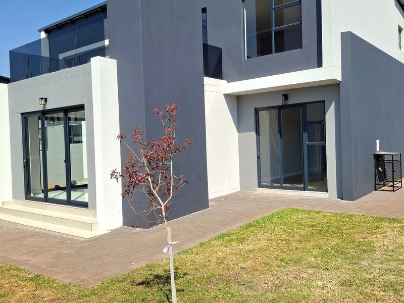 Rear view of a modern two-story house featuring a paved patio, glass sliding doors, and a glass-railed balcony.