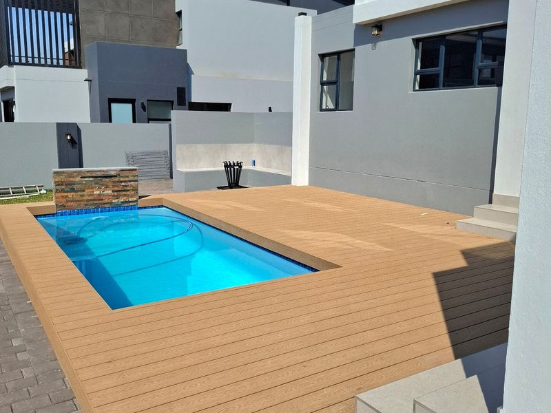 Modern outdoor pool area featuring a rectangular swimming pool, composite decking, and a stone-clad water feature, set against a contemporary grey-toned home exterior.