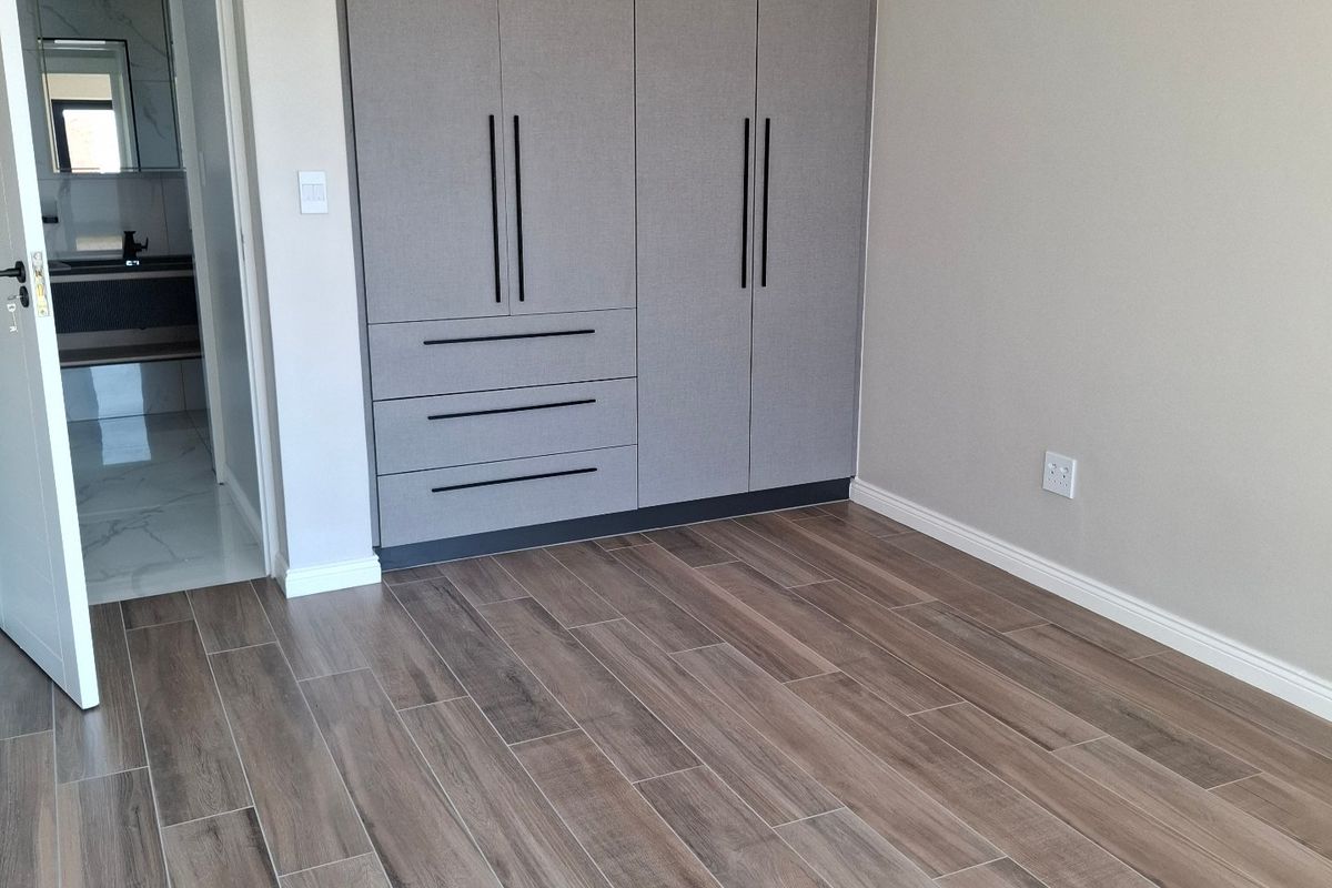 A modern, empty bedroom featuring wood-look tile flooring, built-in grey cupboards, and an adjoining en-suite bathroom.