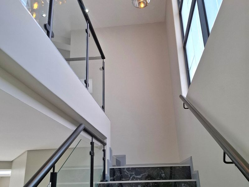 A modern staircase featuring marble steps, a sleek glass balustrade with black metal handrails, and contemporary pendant lighting in a double-volume space.