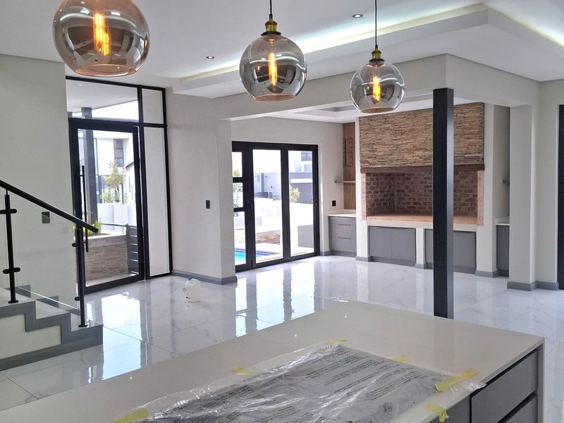 Modern open-plan living area featuring a sleek kitchen island, built-in braai, high-end finishes, and seamless access to the outdoor patio through glass doors.