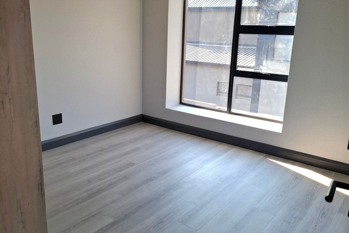 A bright, modern, and empty bedroom featuring light-toned laminate flooring, recessed downlights, and a large aluminium-framed window.