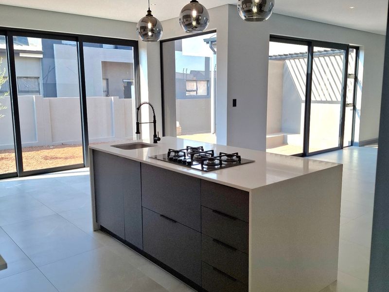 Modern kitchen featuring a central island with a gas hob, undermount sink, and stylish timber slat ceiling detail, opening to an outdoor area.