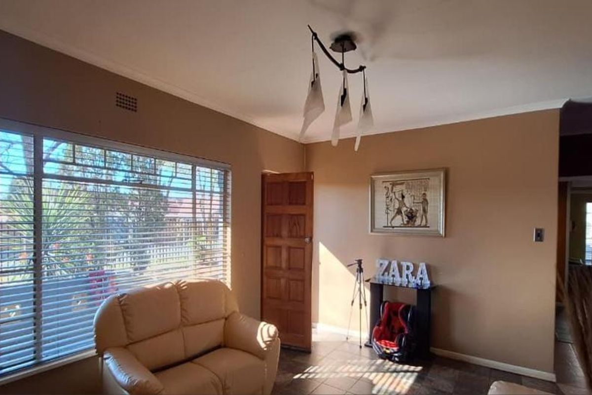 A living room featuring tiled flooring, a ceiling light fixture, and large windows with venetian blinds, providing natural light to the space.