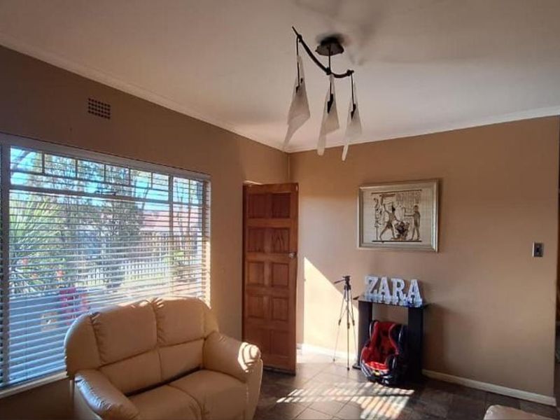 A living room featuring tiled flooring, a ceiling light fixture, and large windows with venetian blinds, providing natural light to the space.