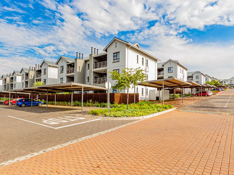 A well-maintained apartment complex featuring paved driveways, covered parking bays, and landscaped garden areas under a clear blue sky.