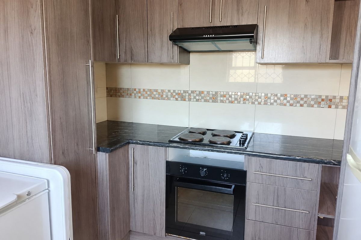 A functional kitchen featuring wood-grain laminate cabinetry, dark stone-look countertops, a built-in electric oven, hob, and a tiled backsplash with decorative accents.