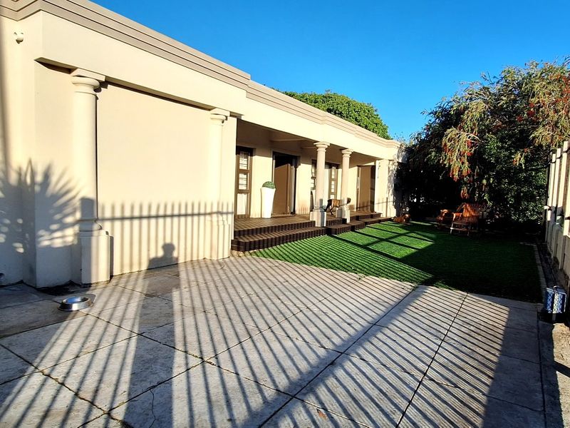 Front exterior view featuring a paved driveway, artificial lawn, covered porch with decorative columns, and a secure metal gate entrance.