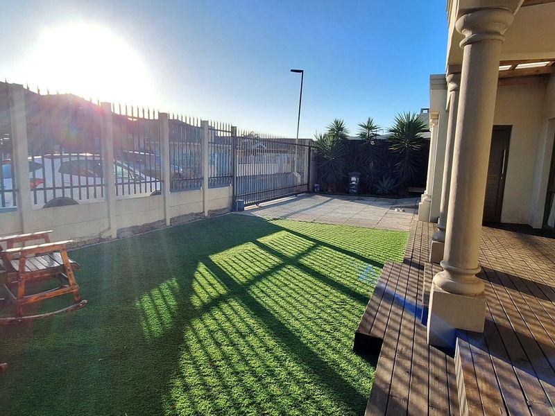 A covered patio area featuring wooden decking and concrete columns, overlooking a small garden space with artificial lawn and perimeter security fencing.