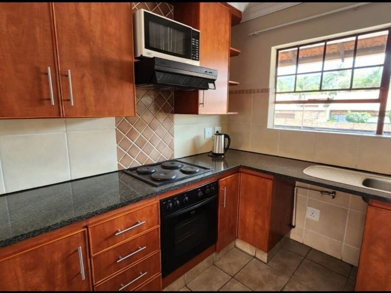 Functional kitchen featuring wood-finish cabinetry, granite countertops, built-in oven, electric hob, and a tiled splashback with natural light from the window.