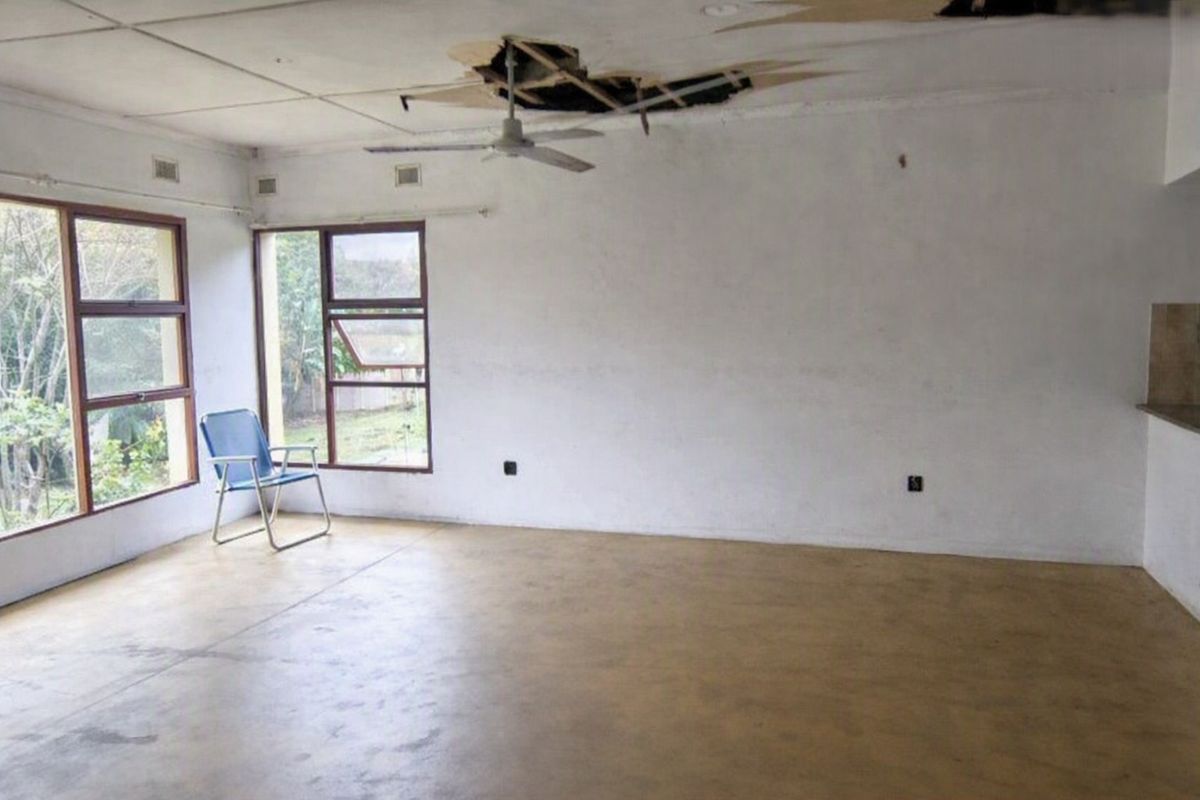 An empty living room featuring large windows and a ceiling fan, currently requiring significant ceiling repairs due to visible structural damage.