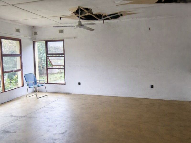 An empty living room featuring large windows and a ceiling fan, currently requiring significant ceiling repairs due to visible structural damage.