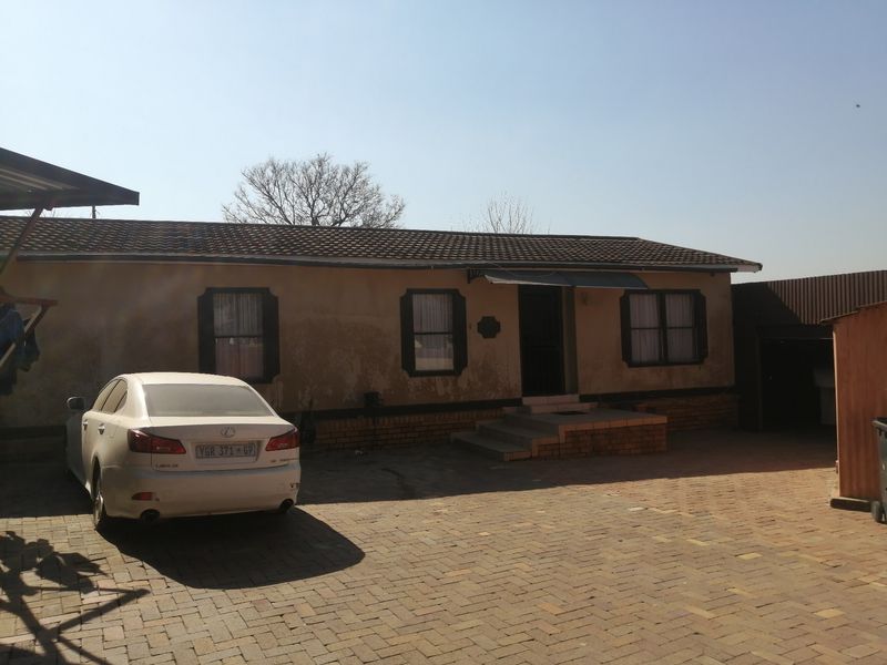 Front elevation of a single-story house featuring a paved driveway, tiled roof, and brick entrance steps, showing signs of exterior wear.